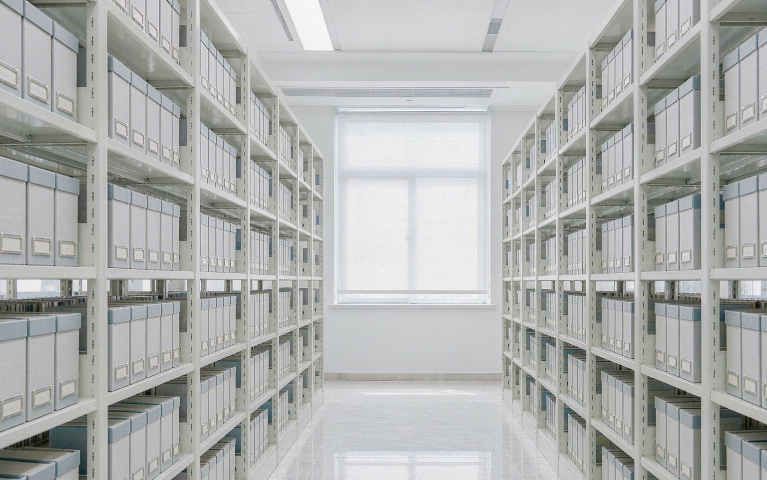 Bright archive shelving aisle lined with uniform grey archive boxes, daylight window at the far end